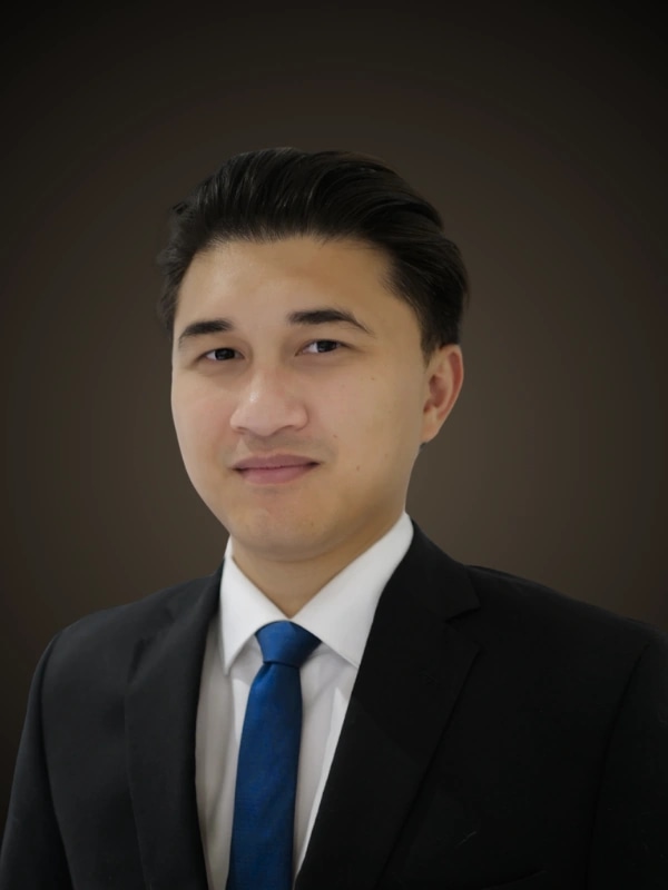 Motta Thong, head and shoulders, in a dark suit, white shirt and blue tie against a dark studio backdrop